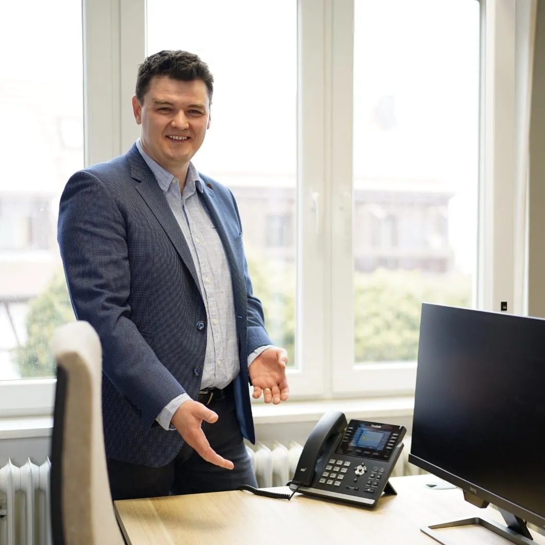 Valerij Schwindt im Sakko zeigt einen hellen Arbeitsplatz mit zwei Monitoren