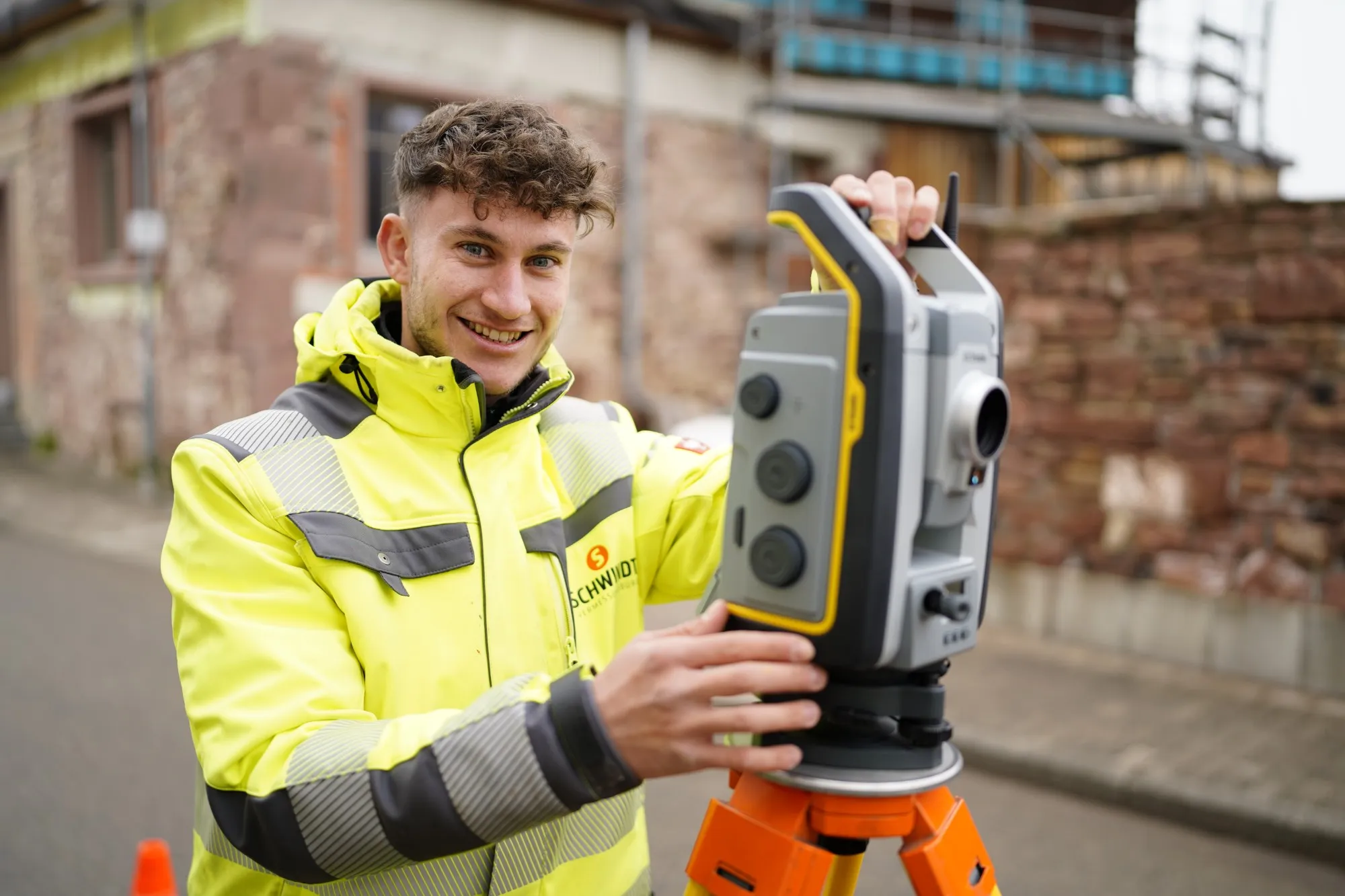 Junger Mitarbeiter in SCHWINDT-Warnjacke lächelt am Tachymeter vor einer Baustelle