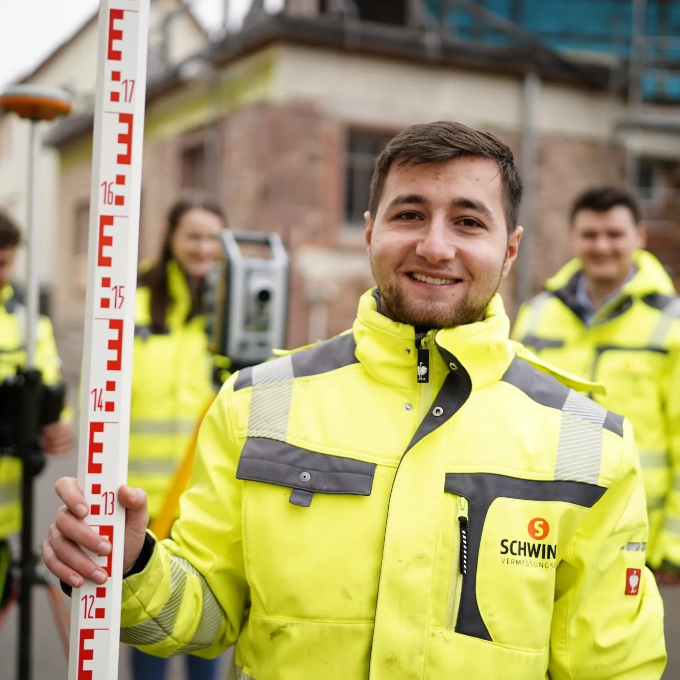Lächelnder Vermessungstechniker in Warnjacke mit Messlatte und Tachymeter, Kolleg*innen im Hintergrund